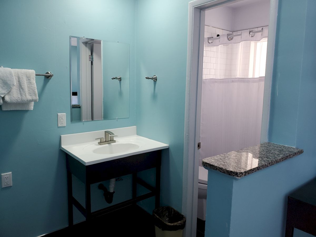 This image shows a bathroom with a sink, a mirror, towel racks, a granite shelf, and an adjoining room with a shower curtain and tiled wall.