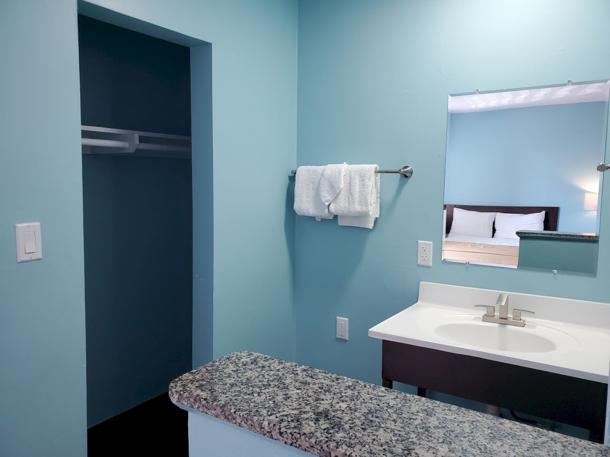 A light blue room with a vanity, a mirror, towels on a rack, and an open closet. A bed with pillows is visible in the reflection on the mirror.