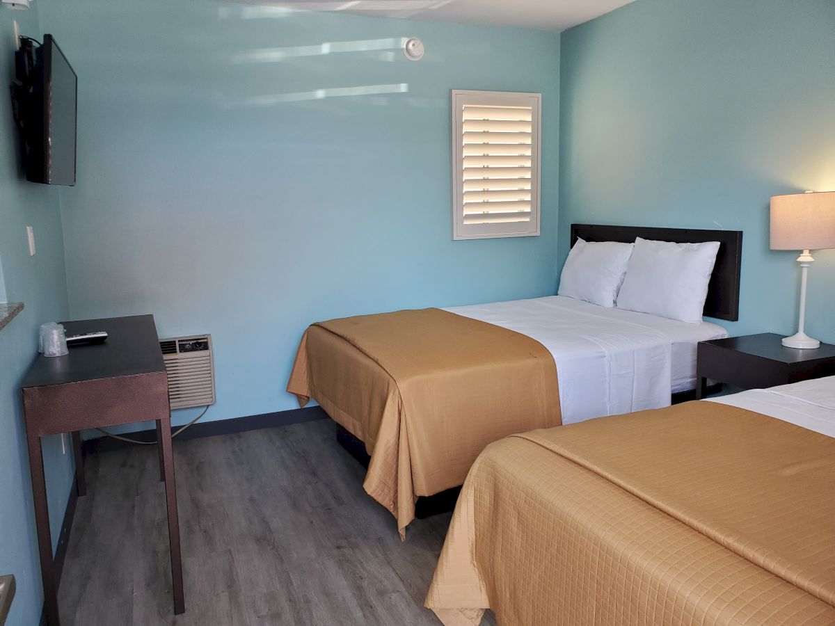 A clean, light blue hotel room with two made beds, a lamp, a small desk, and a wall-mounted TV.